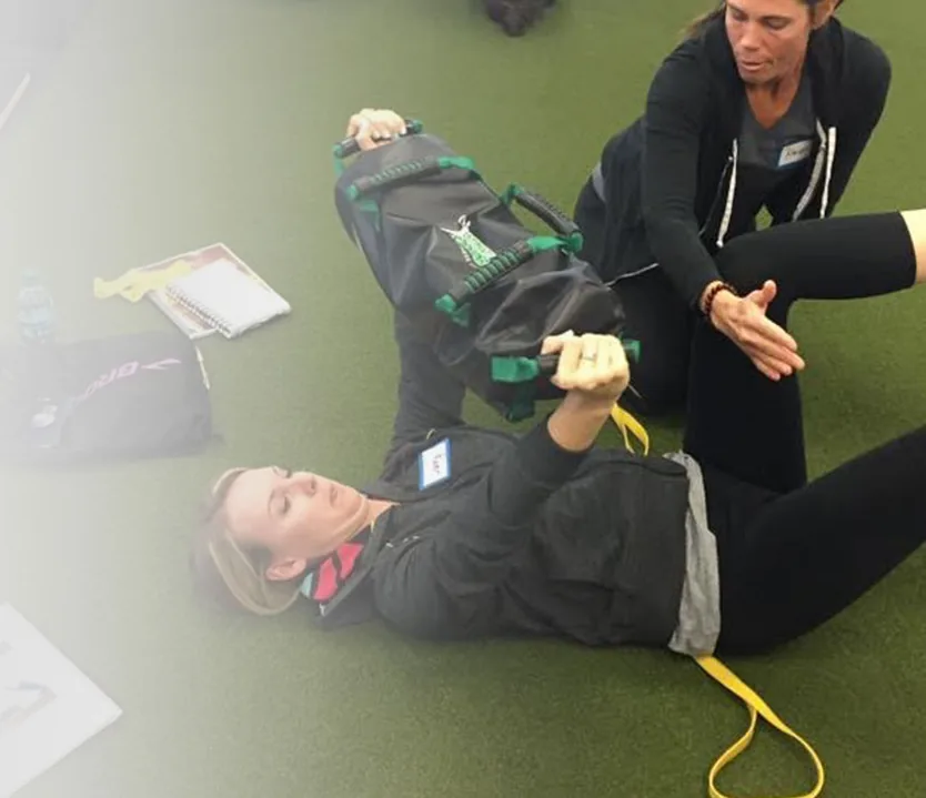 A DVRT instructor coaching a student through a floor drill with a green Ultimate Sandbag during a certification